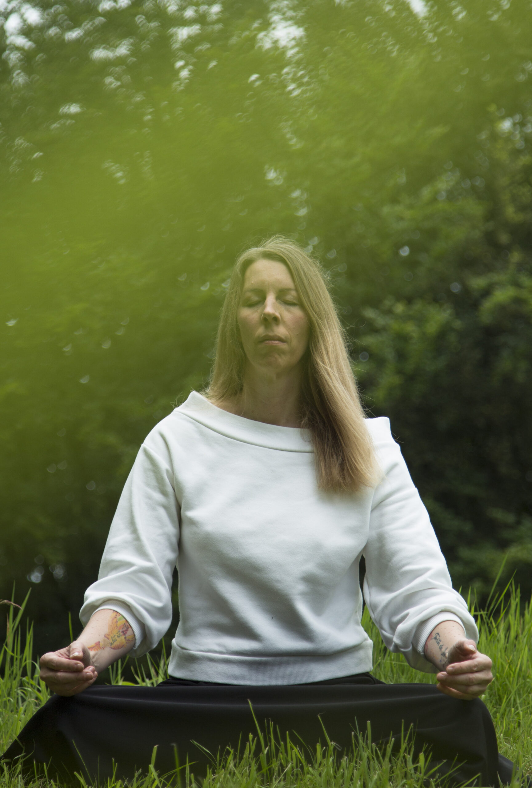 Man sieht Kerstin, eine Frau in ihren 40ern, blonde lange Haare, im Rahmen eines Stille-Retreats tief versunken in Meditation inmitten grüner Natur.