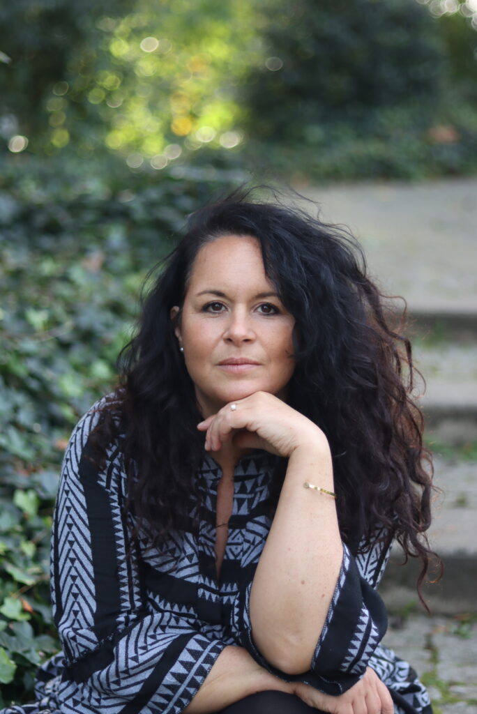 Man sieht eine Frau Mitte 40 vor verschwommenem Naturhintergrund, die mit ruhigem Blick in die Kamera schaut - eine der Team-Mitglieder in der Ausbildung zum Shamanic Practitioner.