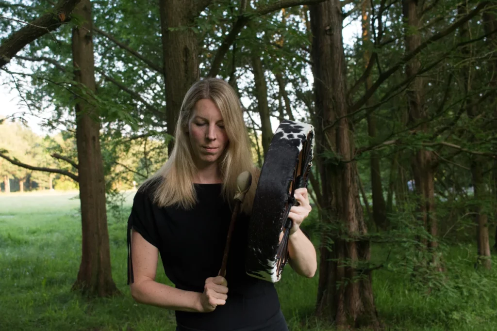 Man sieht Kerstin, eine Frau Mitte 40 mit langen blonden Haaren, in der Natur stehend, im Hintergrund Bäume trommelnd (ihre Trommel ist aus überwiegend schwarzem Fell mit weißen Flecken.