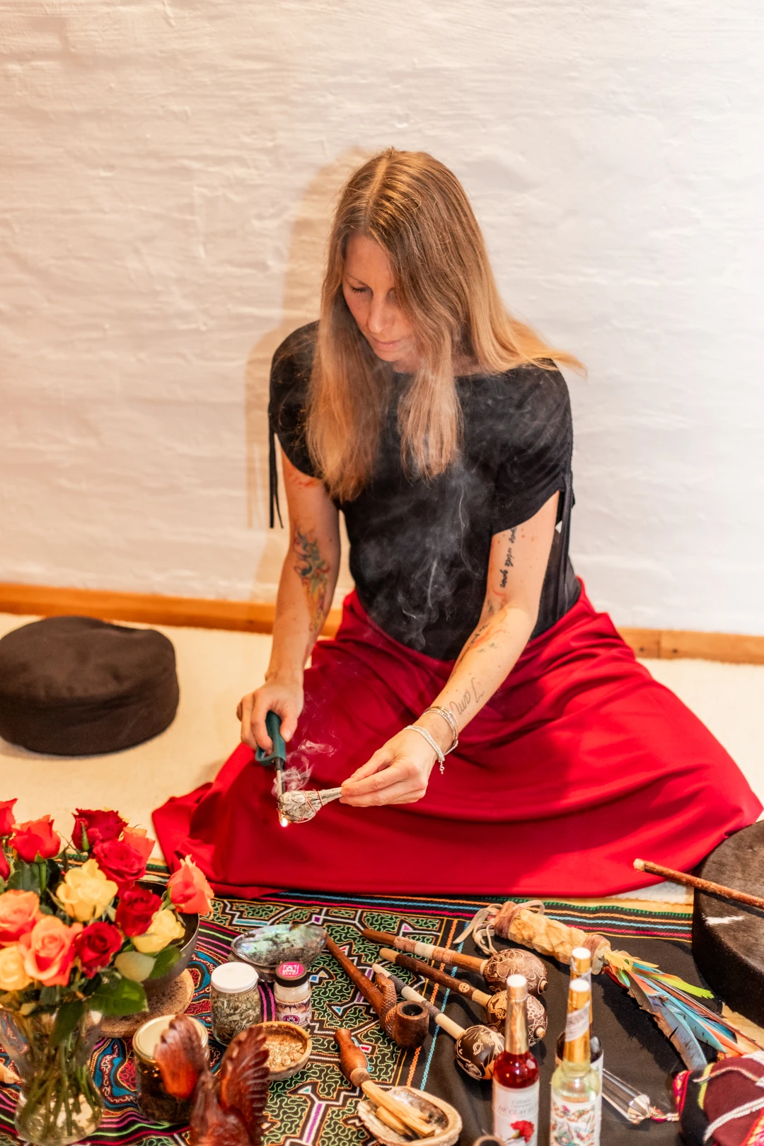 Man sieht Kerstin, eine Frau Mitte 40 mit langen dunkelblonden Haaren und mit langem Rock, vor einem auf dem Boden auf einem Altartuch aufgebauten Altar sitzen. Auf dem Altar befinden sich verschiedenste schamanische Arbeitswerkzeuge, Kerstin entzündet mit einem Feuerzeug ein Salbeibündel, der Rauch steigt auf.