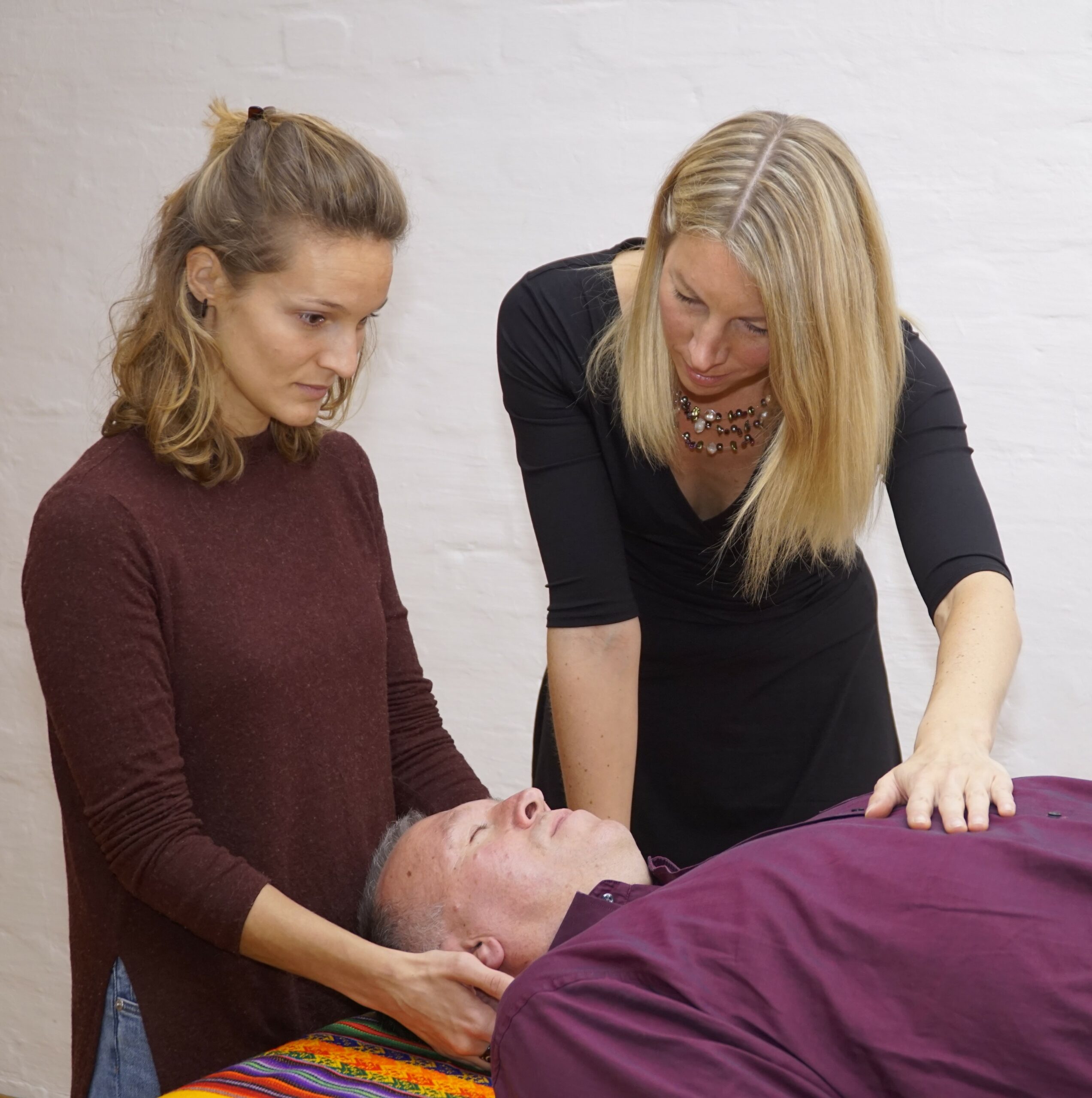 Man sieht im Rahmen der Ausbildung zum/zur Schamanischen BehandlerIn einen Mann auf einer Behandlungsliege und eine junge Frau am Kopfende, die Hände am Hinterkopf des Mannes und der Blick auf seinen Oberkörper gerichtet. Ebenfalls an der Behandlungsliege steht Kerstin, die Lehrerin, mit Blick ins Gesicht des Mannes und einer Hand auf seinem Oberkörper auf Herzhöhe abgelegt.