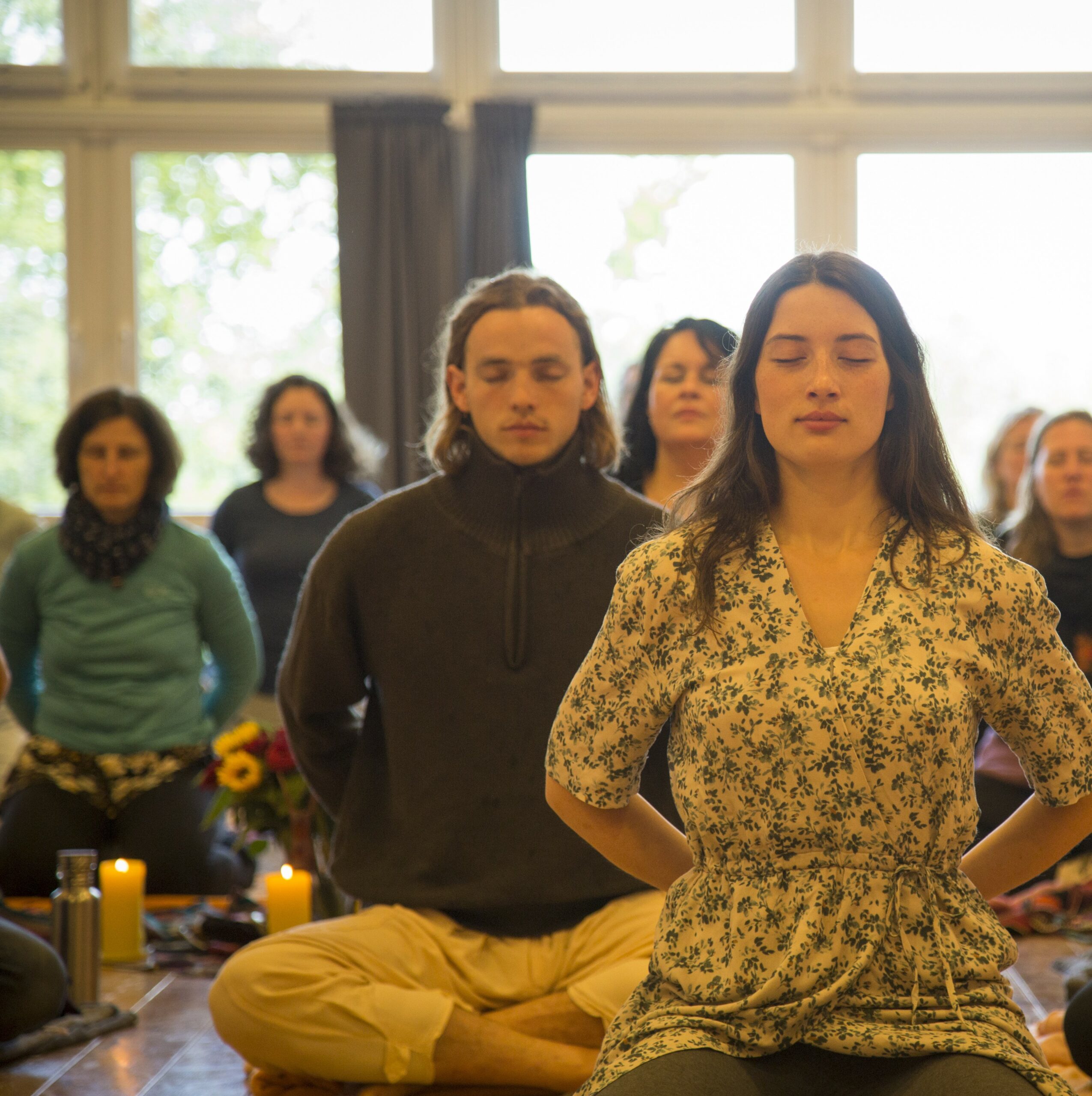 Man sieht eine Gruppe von Menschen verschiedenen Alters und Geschlechts, die mit geschlossenen Augen in die Meditation versunken sind. Auf dem Bild sind ca. 10 - 15 Personen zu sehen bzw. zu erahnen. Im Vordergrund des Bildes eine Frau in ihren 20ern, im Kleid mit langen dunklen Haaren, mit intensiver Ausstrahlung, tief eingetaucht im Rahmen eines schamanischen Stille-Retreats.