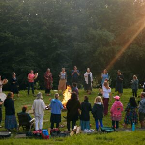 Man sieht eine Gruppe von ca. 35 buntgekleideten Personen verschiedenen Alters auf einer Wiese bzw. Waldlichtung um ein leuchtend brennendes Feuer stehen als Teil des Transformations-Retreats.
