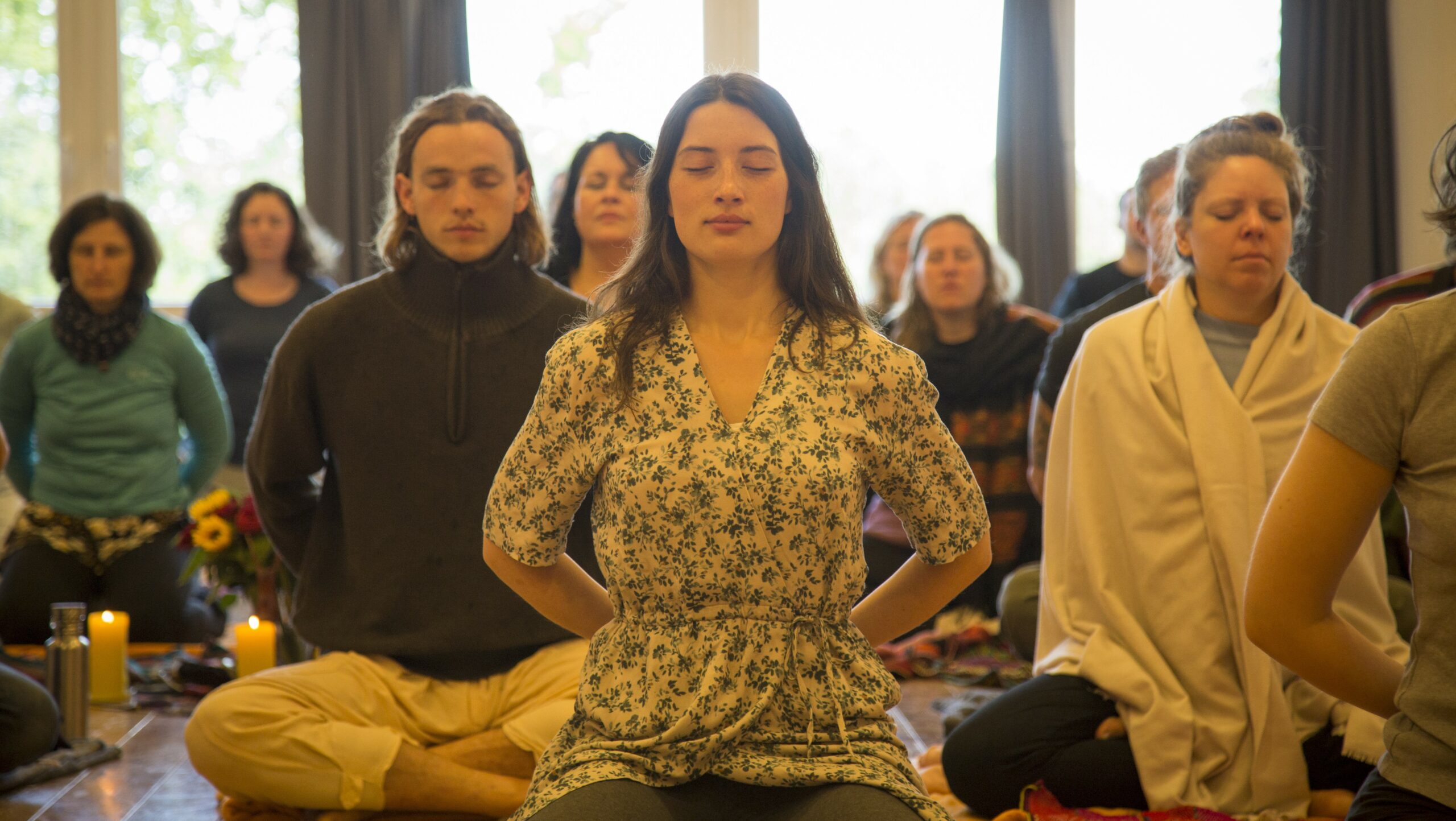 Man sieht eine Gruppe von Menschen verschiedenen Alters und Geschlechts, die mit geschlossenen Augen in die Meditation versunken sind. Auf dem Bild sind ca. 10 - 15 Personen zu sehen bzw. zu erahnen. Im Vordergrund des Bildes eine Frau in ihren 20ern, im Kleid mit langen dunklen Haaren, mit intensiver Ausstrahlung, tief eingetaucht im Rahmen eines Stille-Retreats.