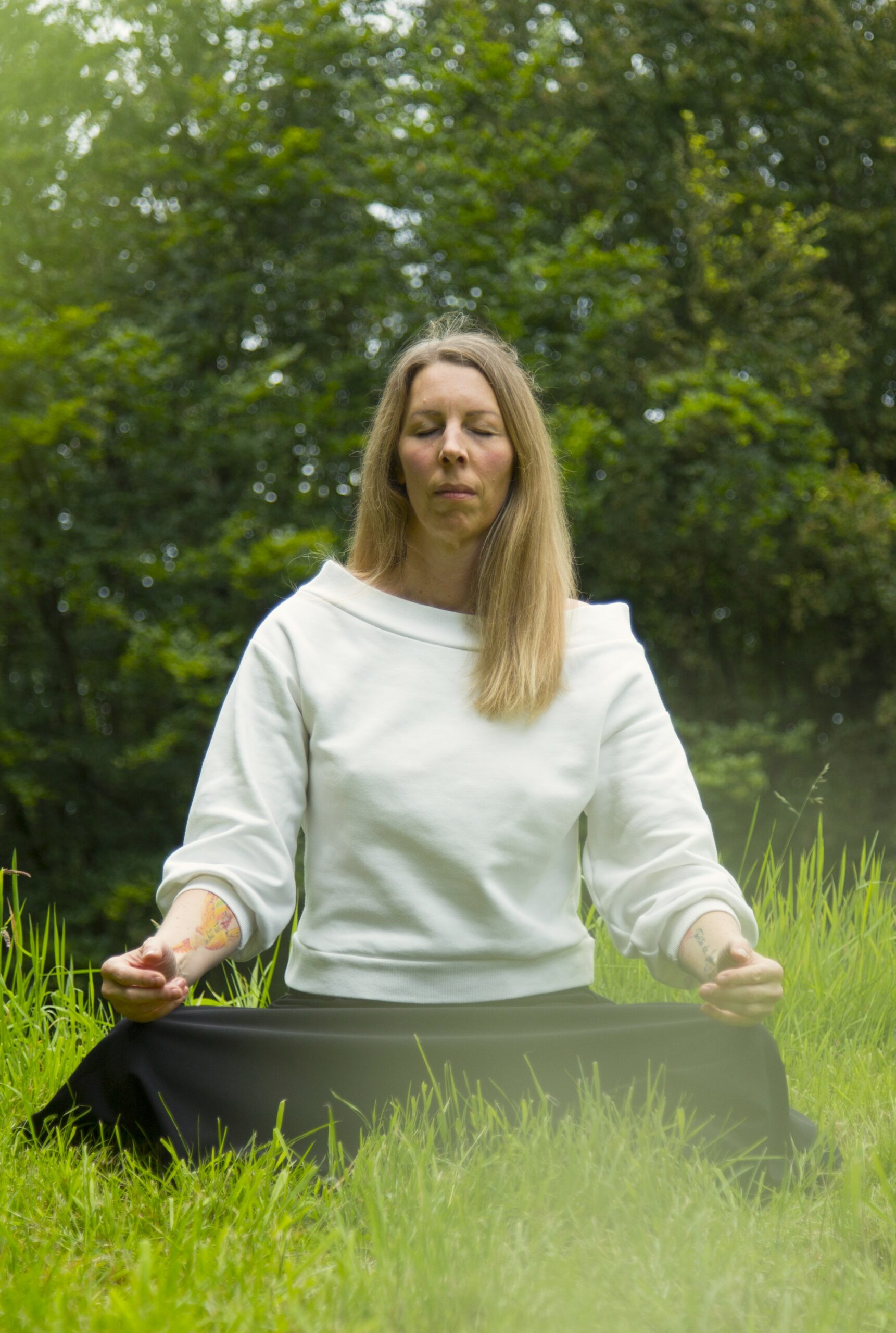 Man sieht Kerstin, eine Frau Mitte 40 mit langen blonden Haaren und langem Rock, im Schneideristz in Meditation versunken, umgeben von Natur.