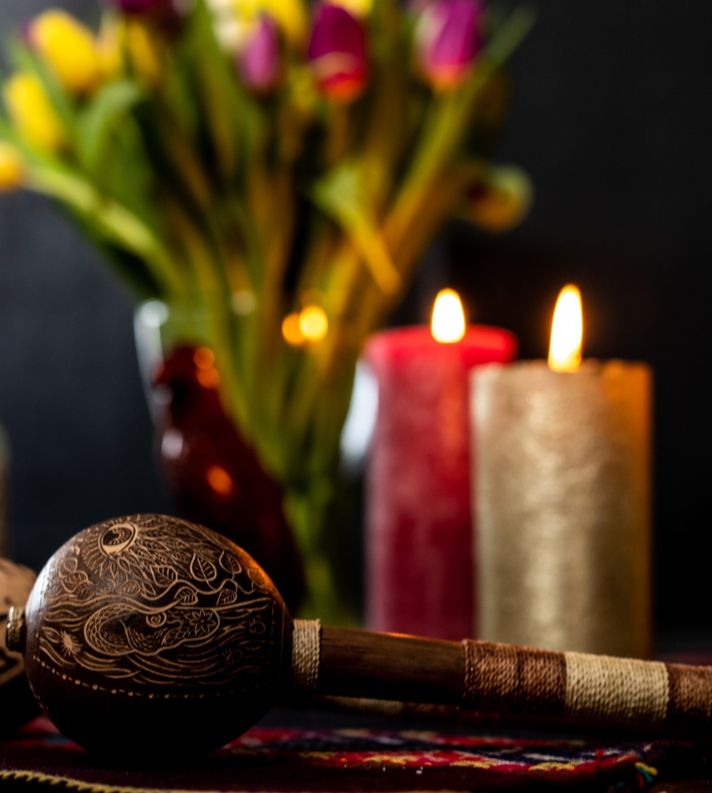 Man sieht vor dunklem Hintergrund in Nahaufnahme auf einem Altartuch eine Rassel mit geschwungenen Lianen-Mustern und einem Auge darauf sowie im Hintergrund brennende Kerzen und Tulpen in einer Vase.