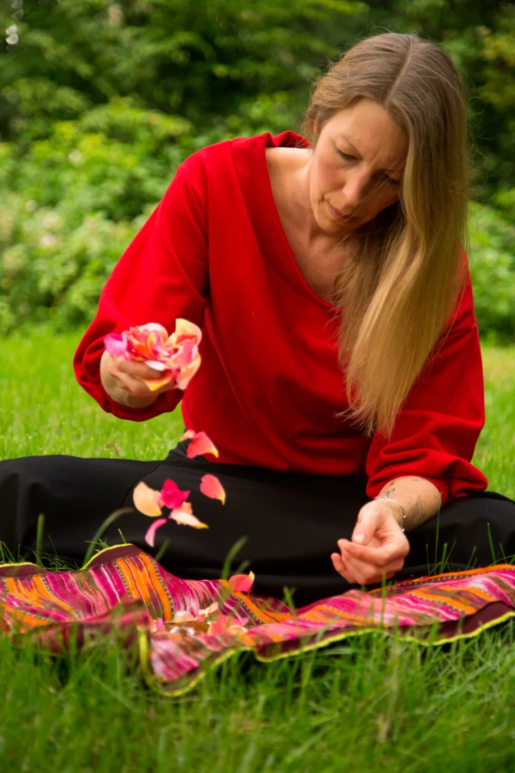 Man sieht Kerstin, eine Frau Mitte 40 mit blonden langen Haaren. Sie sitzt auf einer Wiese und lässt mit Sorgfalt Rosenblütenblätter auf ihr Medizinbündel fallen.