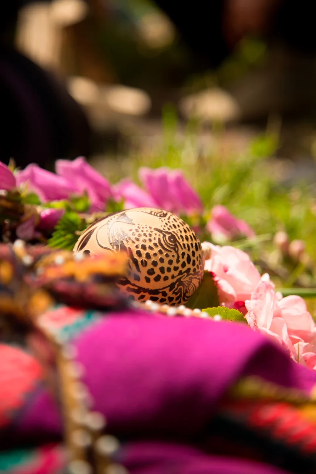 Man sieht im Zentrum des Bildes eine Rassel mit eingeschnitztem Jaguarkopf, drumherum unscharf ein buntes Medizinbündel sowie viele rosa und pinke Blüten und Blumen, im Hintergrund Gras.