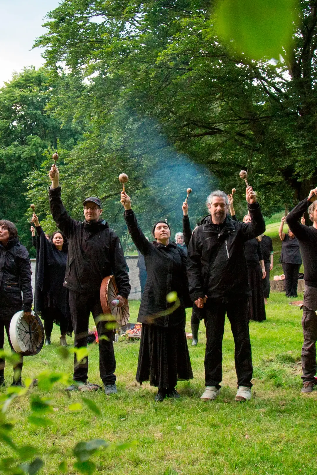 Man sieht eine Gruppe mehrerer Personen in den 30ern und 40ern, schwarz gekleidet, mit Rasseln und zum Teil mit Trommeln in ihren Händen und die Hand mit der Rassel zum Himmel erhoben, in grüner Natur umgeben von Bäumen, im Hintergrund ein noch brennendes Feuer in einer Feuerstelle, die sich im Rahmen eines schamanischen Transformations-Retreats zum Abschluss eines Rituals bei den sie unterstützenden Kräften der geistigen Welt bedanken.