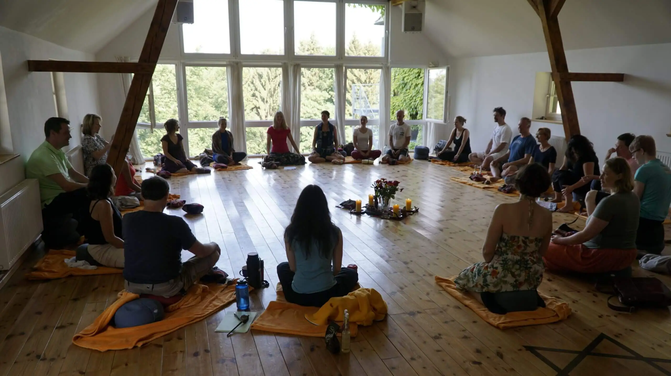 Man sieht eine Gruppe von ca. 30 Personen in einem Sitzkreis mit Sitzkissen in einem großen, hellen Seminarraum mit einem Altar u.a. mit Blumen und Kerzen im Zentrum des Raumes.