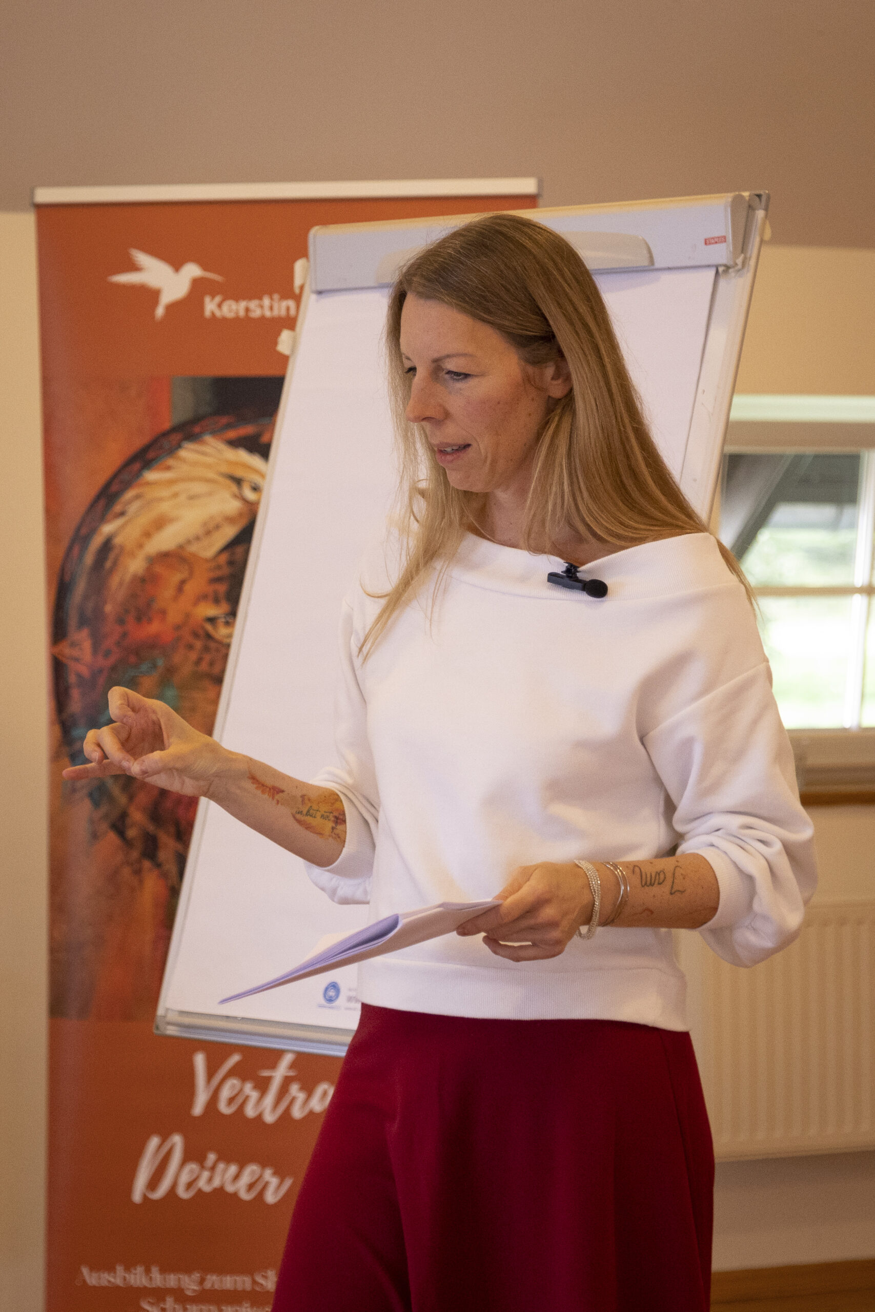 Man sieht eine Frau in ihren Vierzigern, die vor einem Flipchart steht, mit Notizen in der Hand, und spricht.