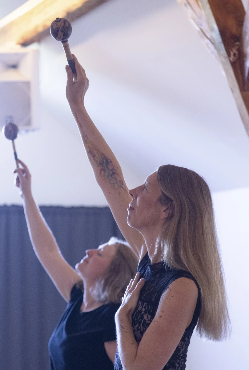 Man sieht zwei Frauen in einem Seminarraum, die jede in enier Hand eine Rassel halten und diese gen Himmel gestreckt haben - eine mit offenen Augen gen Himmel und Hand auf dem Herzen, eine mit geschlossenen Augen - die Kraft der Manifestation.