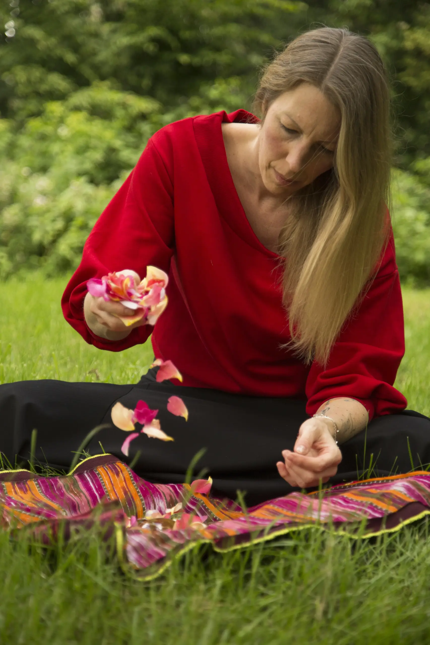 Man sieht eine Frau in ihren Vierzigern inmitten grüner Natur, die Rosenblütenblätter auf eine Tuch fallen lässt - die Geheimnisse der Manifestation.