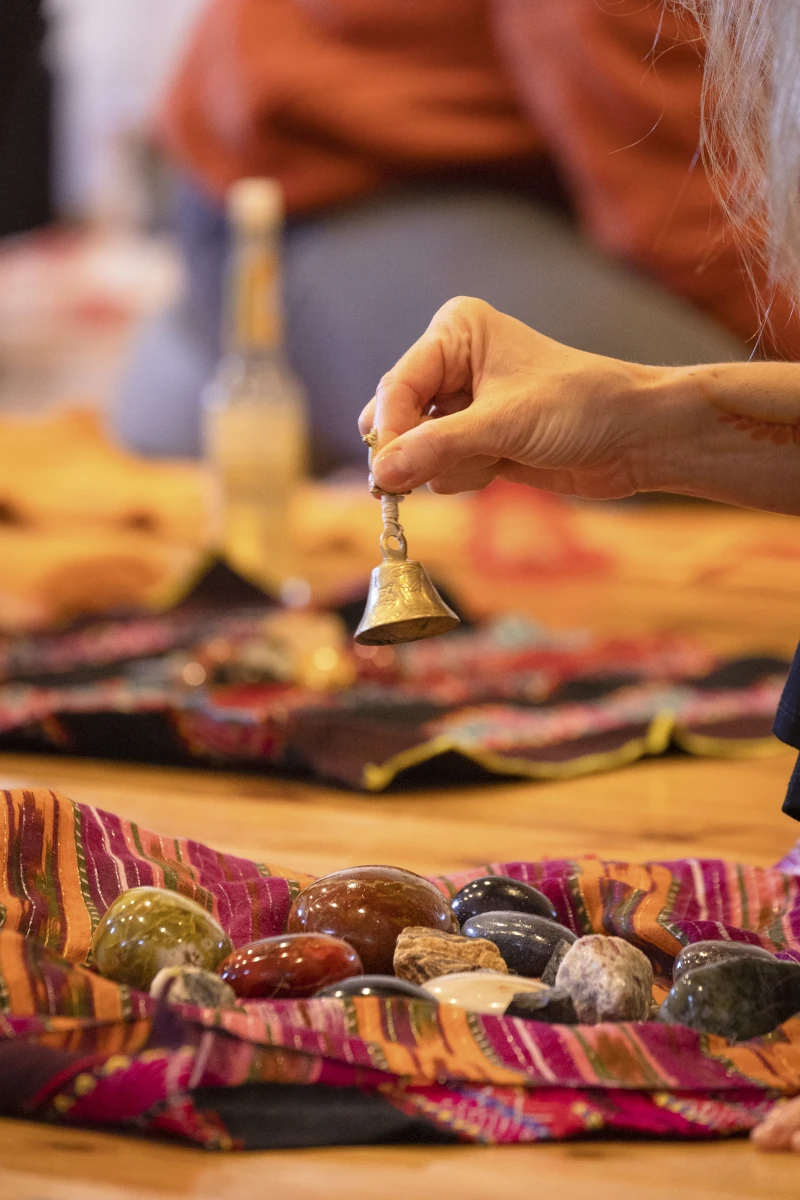 Man sieht in Nahaufnahme eine Hand, die ein kleines Glöckchen über bunten Medizinsteinen läutet - die Geheimnisse der Manifestation.