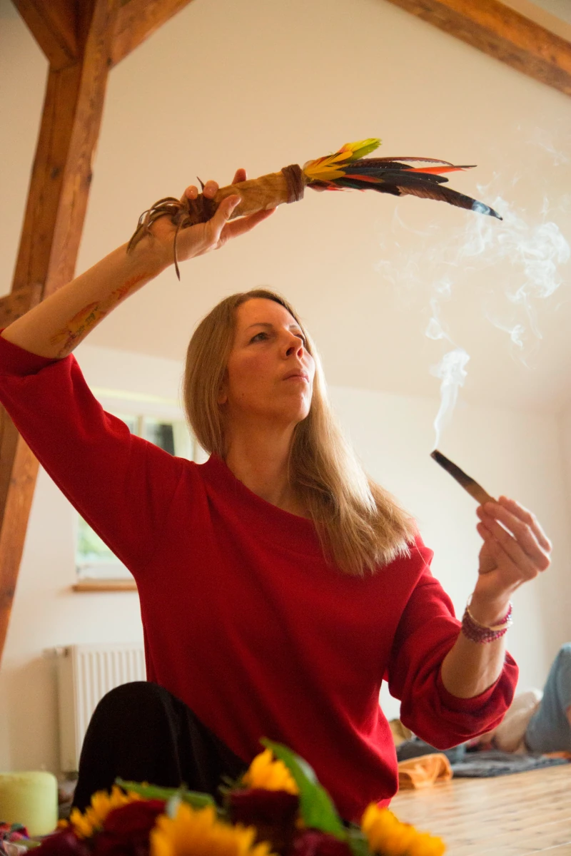 Man sieht eine Frau in ihren Vierzigern, die in einer Hand einen Palo Santo-Stick hält, von dem Rauch aufsteigt, und in der anderen Hand ein Bündel bunter Federn. Die Frau ist konzentriert auf den Rauch - Räuchern im Schamanismus.