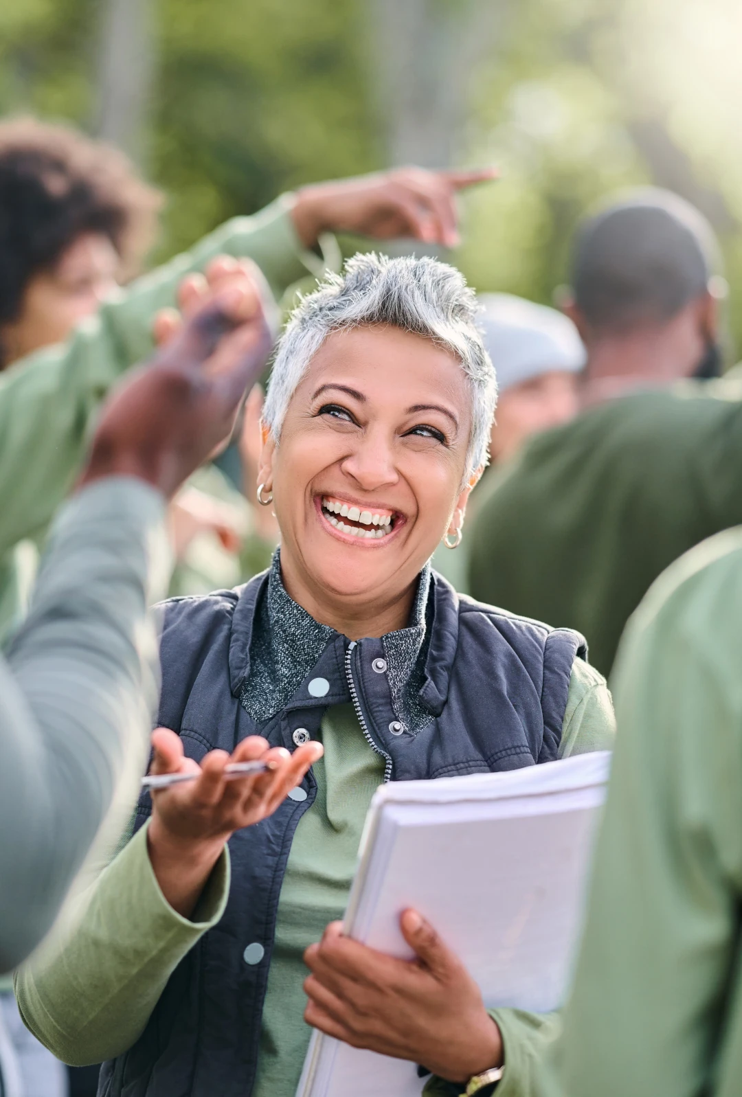 Man sieht eine Frau etwa in ihren Fünfzigern in einer Gruppe von Menschen, die mit Block und Stift in der Hand lachend im Gespräch mit jemandem ist - Beitrag leisten und persönliche Bedeutung als zwei unserer Grundbedürfnisse.