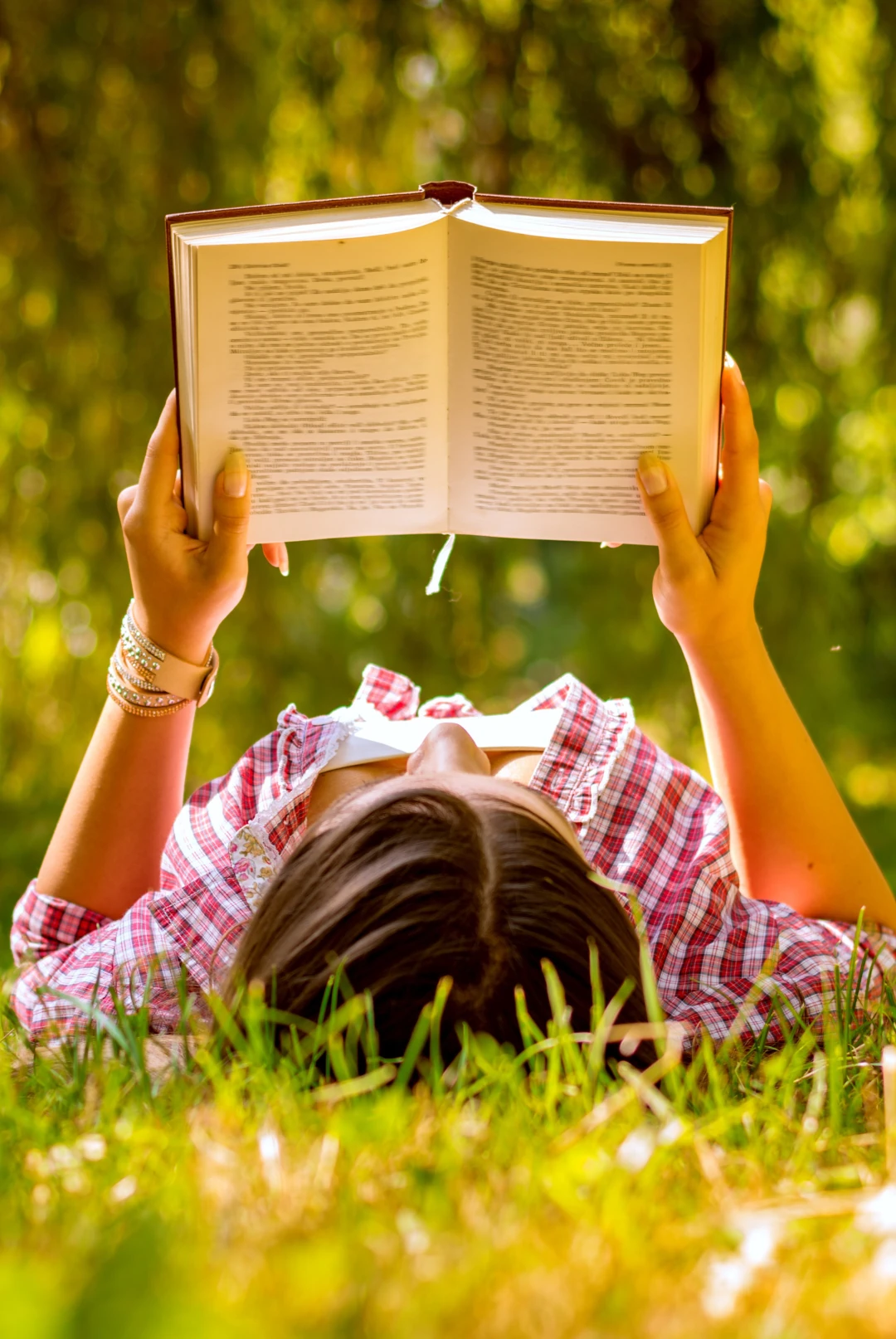 Man sieht vom Kopfende her fotografiert eine junge Frau, die auf dem Rücken auf einer grünen Wiese liegt und ein Buch in ihren Händen hält - Wachstum als eines unserer Grundbedürfnisse.