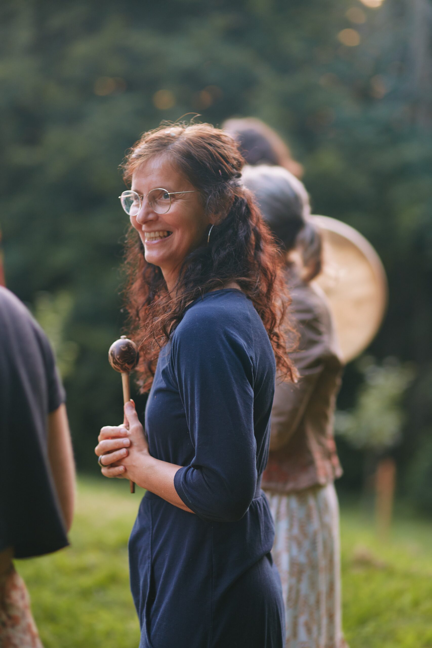 Hier sieht man im Rahmen des Weiblichkeits-Retreats eine Frau Anfang 50 mit strahlendem Gesichtsausdruck draußen in der Natur, mit einer Rassel in der Hand; verschwommen sind im Hintergrund weitere Frauen zu erkennen.