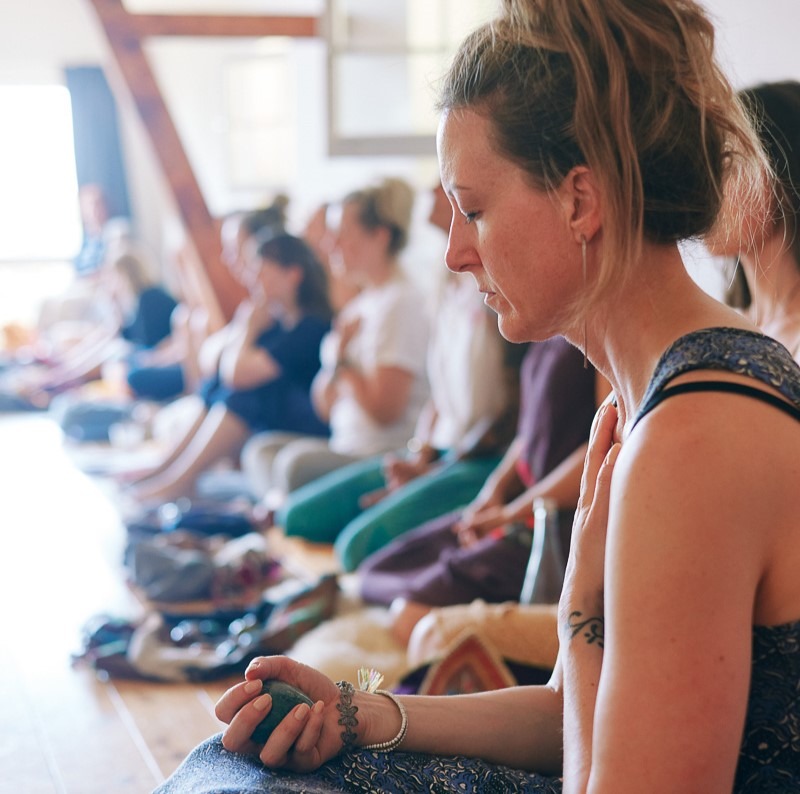 Man sieht im Vordergrund des Bildes eine Frau um die 40, im Profil fotografiert in Nahaufnahme, mit geschlossenen Augen und mit einer Hand auf dem Herzen versunken in einen inneren Prozess, im Hintergrund verschwommen weitere Frauen ebenfalls mit geschlossenen Augen als Teil des Stille-Retreats.