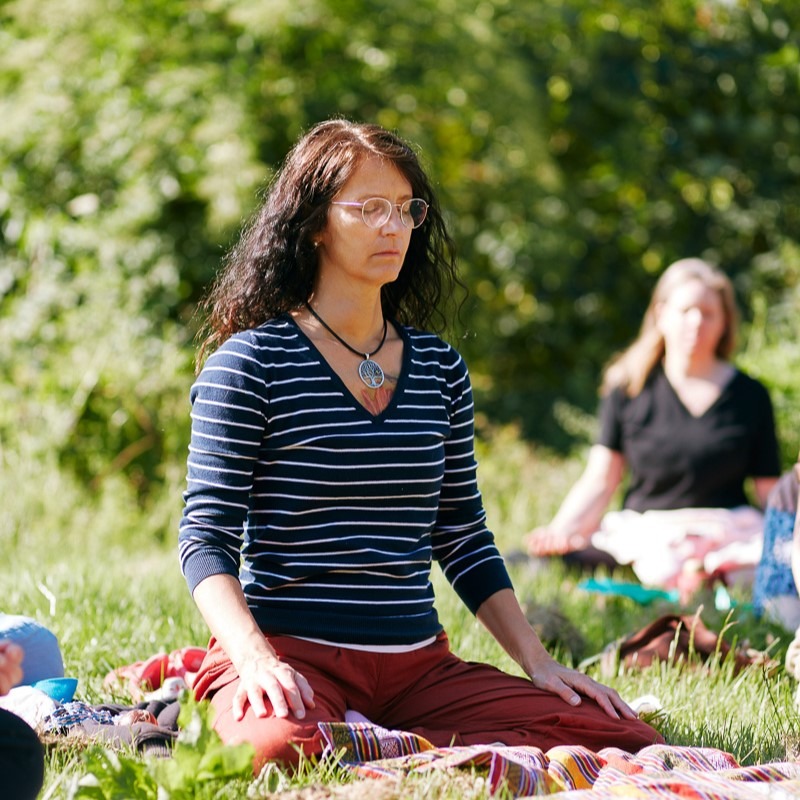 Man sieht im Rahmen des Stille-Retreats eine Frau in ihren 50ern auf einer Wiese in der Sonne mit geschlossenen Augen meditierend, hinter ihr weitere Frauen, ebenfalls meditierend.