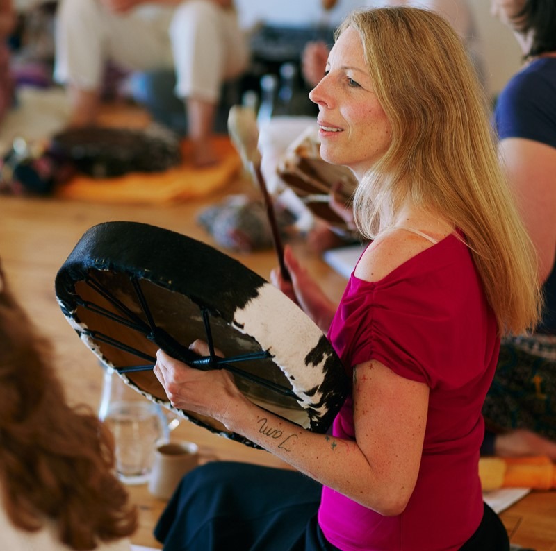 Man sieht eine Frau in ihren 40ern, die im Rahmen des Workshops Schamanisches Träumen auf einer Trommel spielt, um sie herum verschwommen weitere Personen.