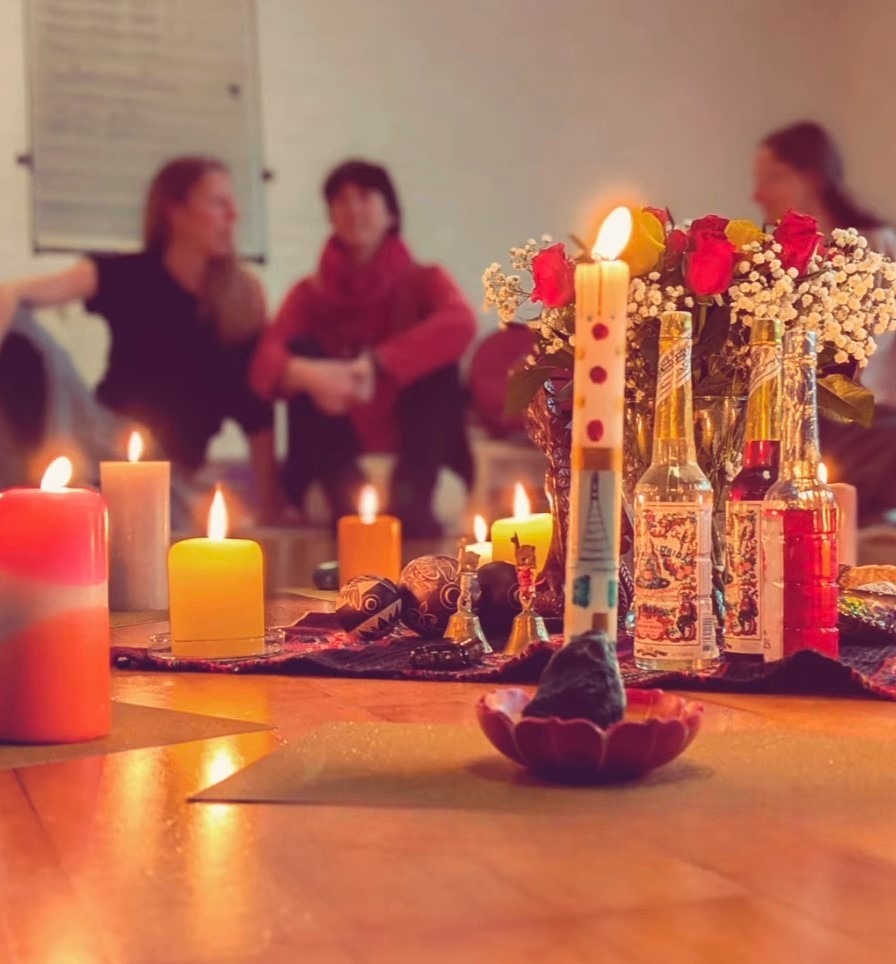 Hier sieht man im Rahmen eines Lebensglück-Workshops im Vordergrund des Bildes einen Altar mit Blumen, brennenden Kerzen und schamanischen Arbeitswerkzeugen sowie weitere brennende Kerzen um den Altar herum; im Hintergrund, etwas verschwommen, sieht man drei Frauen in ihren 40ern, die auf dem Boden sitzen und sich unterhalten.