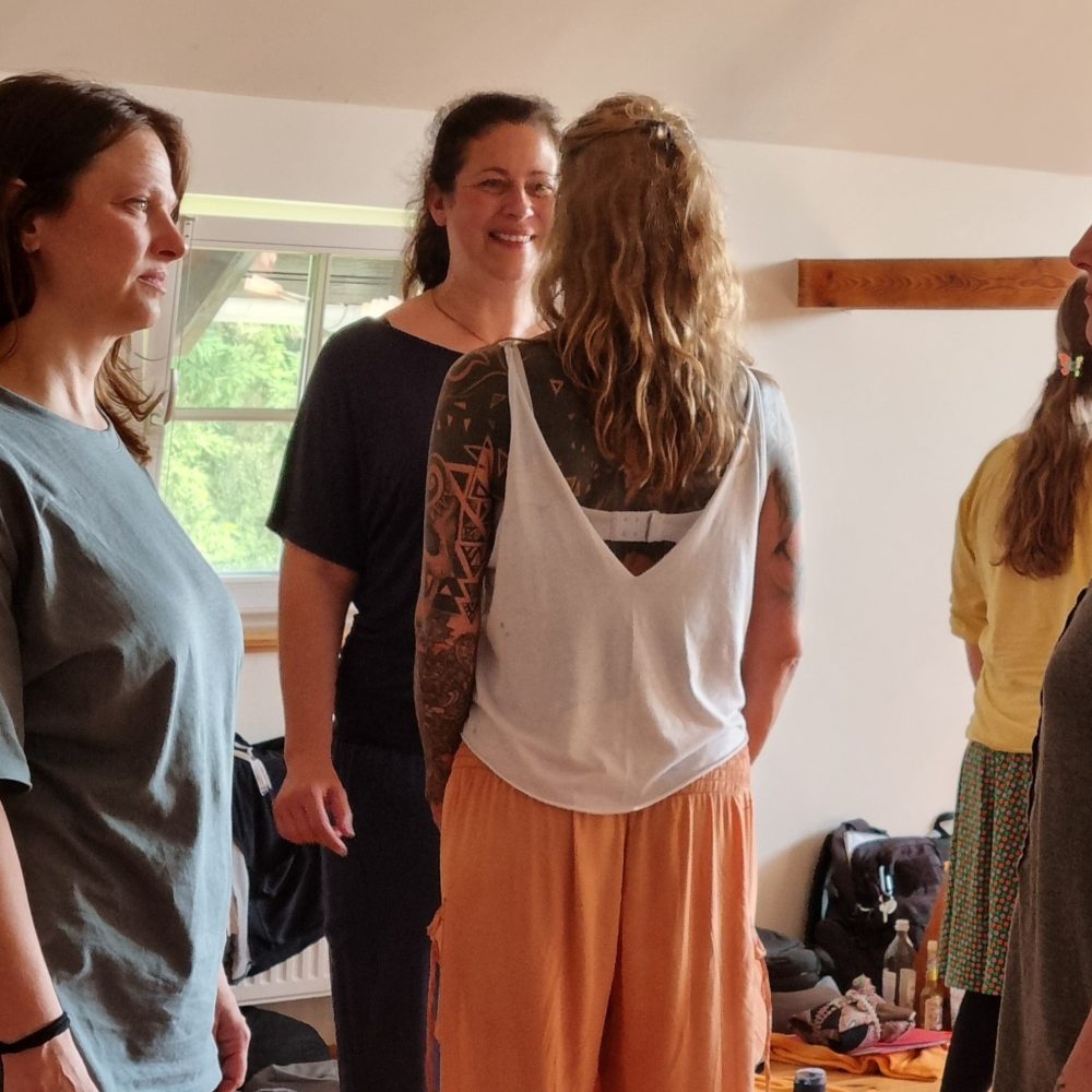 Hier sind man im Rahmen eines der Lebensglück-Workshops zwei Frauen, die sich gegenüberstehen und sich in die Augen schauen; weitere Frauen ebenfalls in Begegnungen am Rande des Bildes.