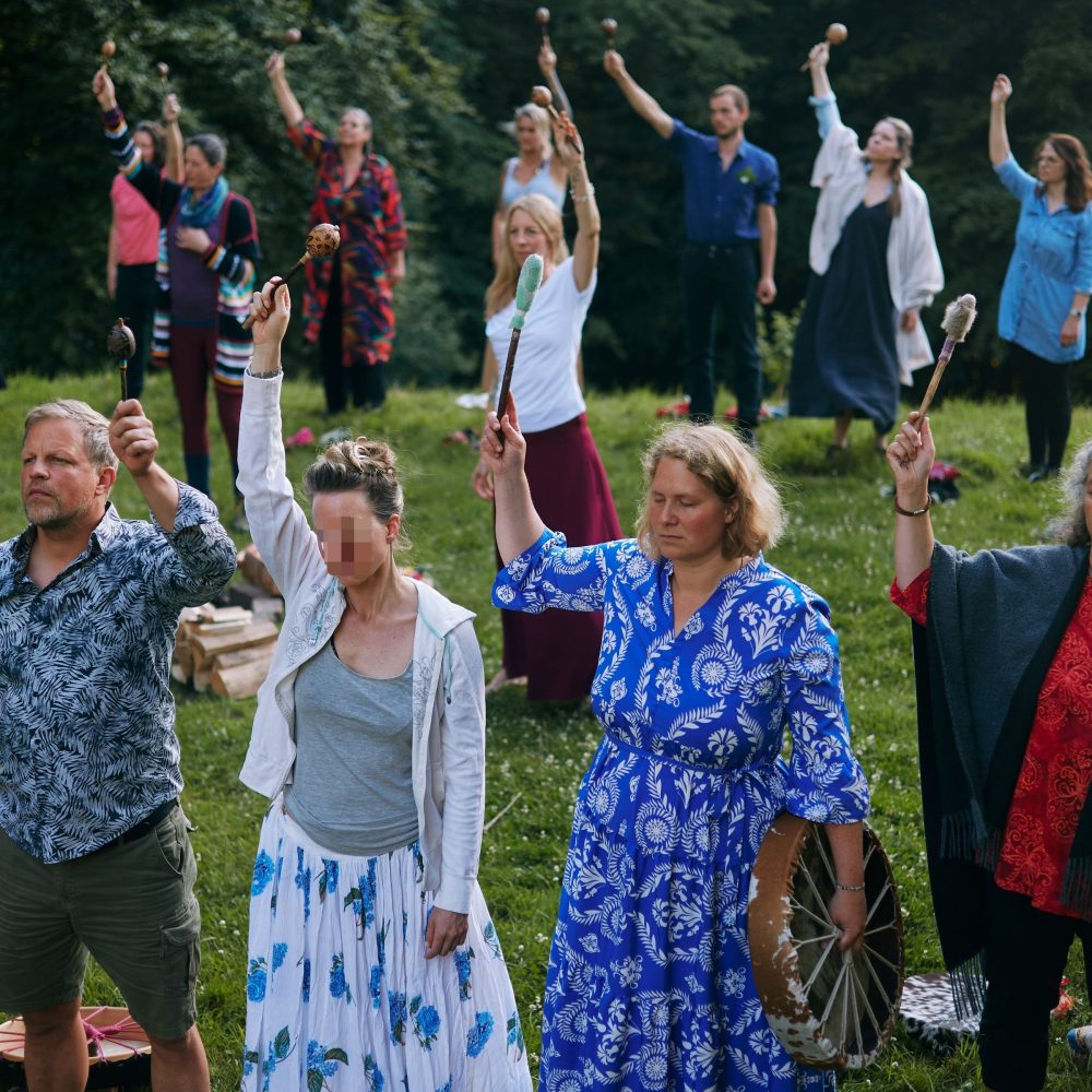 Hier sieht man im Rahmen des Transformations-Retreats eine Gruppe buntgekleideter Menschen um eine noch nicht entzündete Feuerstelle herum; alle stehen in eine Richtung gewandt.