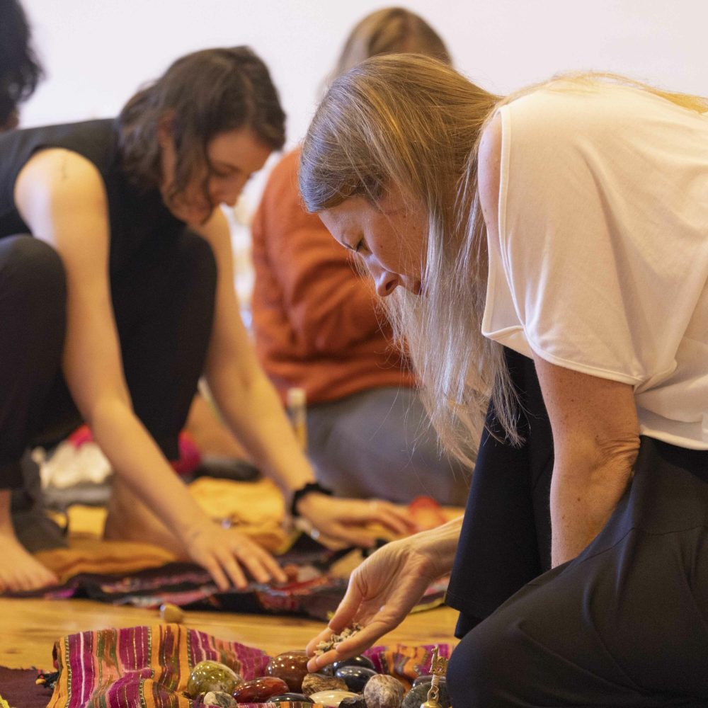 Man sieht im Rahmen der Ausbildung zum Shamanic Practitioner im Vordergrund den Ausschnitt einer Frau, die sich über ihr geöffnetes Medizinbündel beugt und aus ihrer Handfläche Maiskörner in das Bündel rieseln lässt. Im Hintergrund weitere Personen, die ebenfalls mit ihrem eigenen Medizinbündel beschäftigt sind.