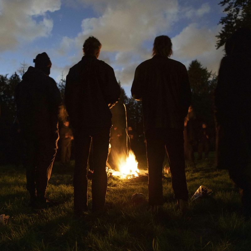 Man sieht im Rahmen einer schamanischen Feuerzeremonie eine Gruppe von Menschen in der Dunkelheit um ein leuchtendes Feuer stehen.