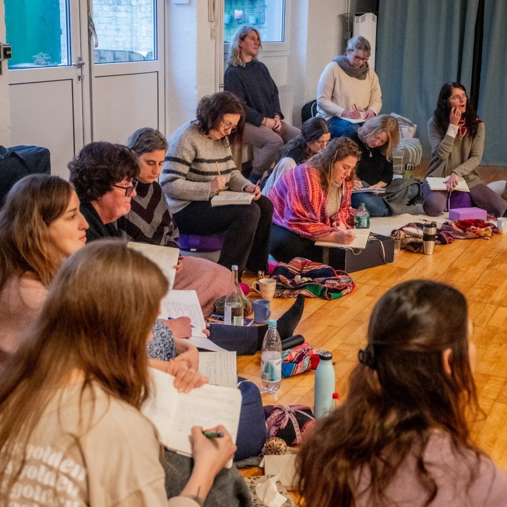 Man sieht eine Gruppe von ca. 15 Frauen verschiedenen Alters, die in der Ausbildung zur Schamanischen Behandlerin in einem Halbkreis sitzen und (bei einer Demo) intensiv mitschreiben.