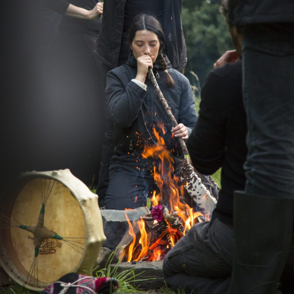 Man sieht eine Frau in ihren 20ern, die an einem brennenden Feuer mit geschlossenen Augen und versunkenem Gesichtsausdruck in einen großen Stock hineinbläst, den sie in ihren Händen hält. Drumherum sieht man verschwommen weitere Personen am Feuer hockend - Feuerrituale sind Teil der Ausbildung zum Shamanic Practitioner.