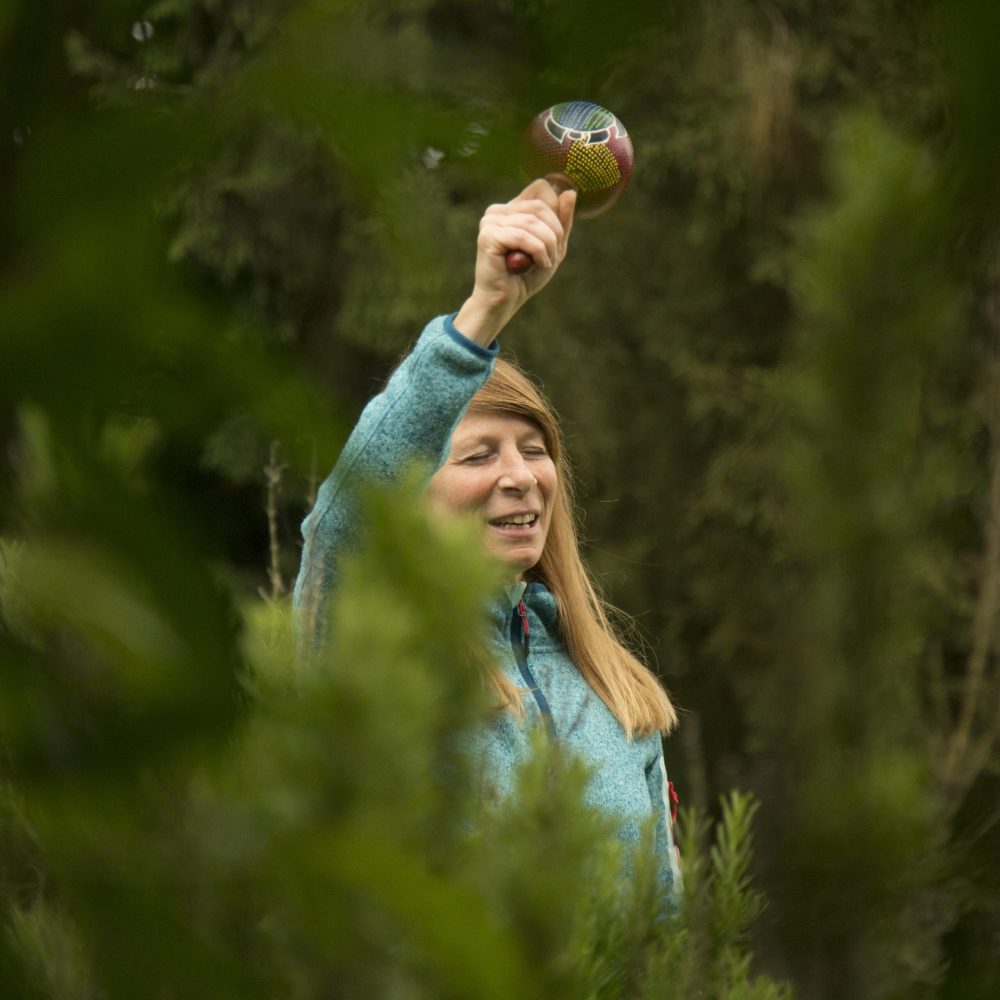 Man sieht im Rahmen der Ausbildung zum Shamanic Practitioner zwischen den Bäumen hervorlugend eine Frau, die mit geschlossenen Augen und strahlendem Gesichtsausdruck eine Rassel gen Himmel hält.