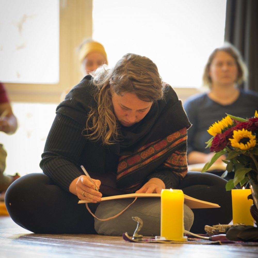 Man sieht im Rahmen der Ausbildung zum Shamanic Practitioner eine Frau in ihren 30ern auf dem Boden sitzend, vor sich auf einem Sitzkissten ihr geöffnetes Notzibuch, in das sie konzentriert hineinschreibt; neben ihr Kerzen und Blumen als Ausschnitt des Altars, im Hintergrund verschwommen weitere Personen.