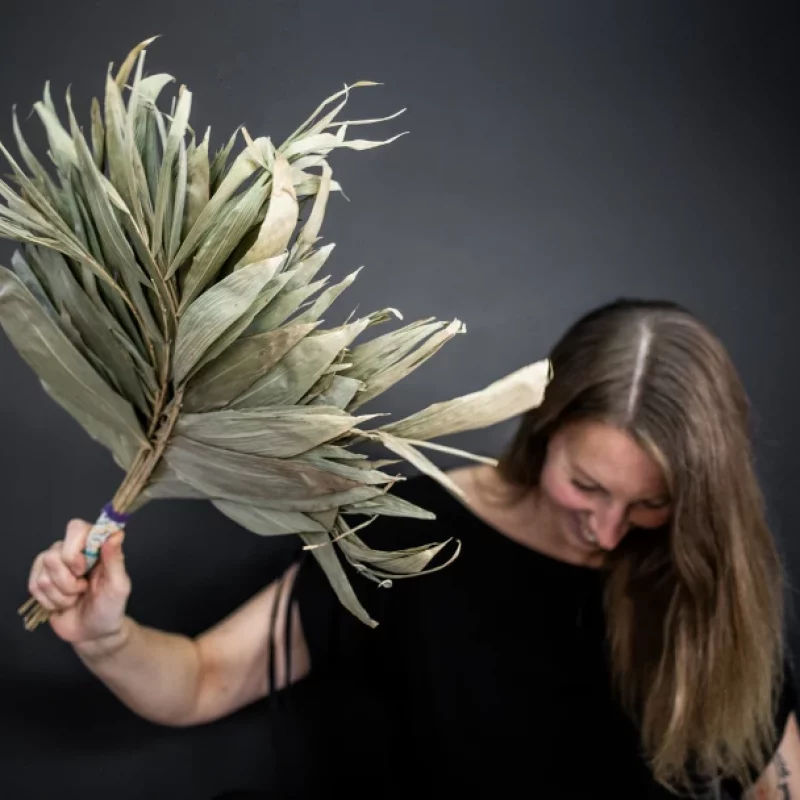 Man sieht Kerstin, eine Frau Mitte 40 mit langen blonden Haaren vor schwarzem Hintergrund, die im Rahmen einer Behandlung mit Schamanischer Energiemedizin ein großes Blätterbündel in einer Hand hält und schüttelt.