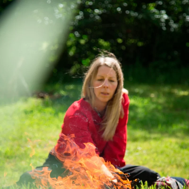 Man sieht im Vordergrund des Bildes ein Feuer, am Feuer sitzt im Schneidersitz eine Frau Mitte 40, den Blick auf das Feuer gerichtet und eine Hand zum Feuer ausgestreckt; Teil einer schamanischen Feuerzeremonie.