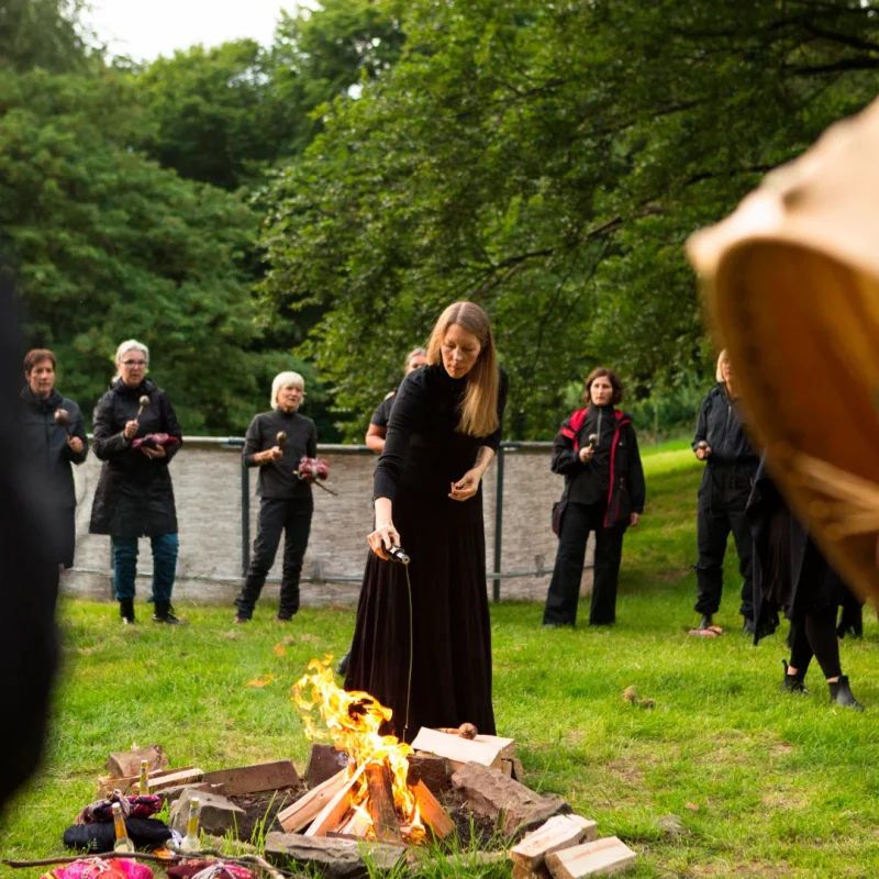 Man sieht, wie eine schwarzgekleidete Frau Mitte 40 mit langem Rock in einer schamanischen Feuerzeremonie am Feuer stehend Öl auf das brennende Feuer gießt; im Hintergrund der Ausschnitt eines Kreises vieler Personen und Bäume.