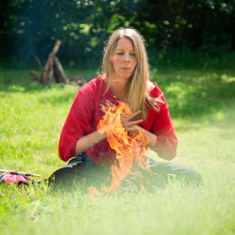 Man sieht im Vordergrund des Bildes ein Feuer, am Feuer sitzt im Schneidersitz eine Frau Mitte 40, den Blick auf das Feuer gerichtet und die Hände vor dem Herzen; Teil einer schamanischen Feuerzeremonie.