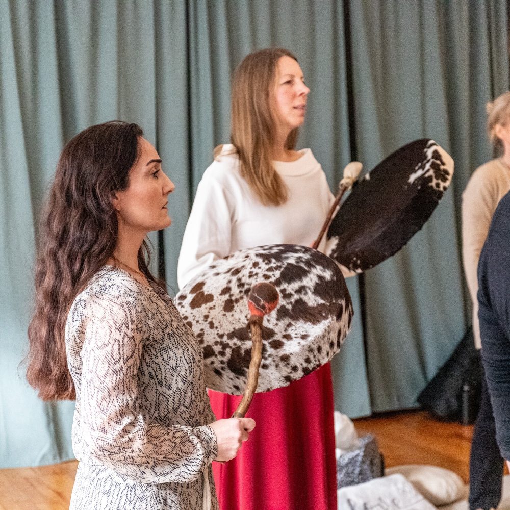 Man sieht im Rahmen eines Schamanische Gesänge-Workshops zwei Frauen in ihren 40ern, die stehend auf ihre Trommeln schlagen und singen; am Rand sind Ausschnitte weiterer Personen erkennbar.