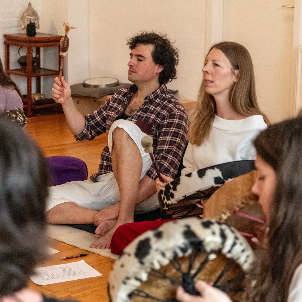 Man sieht im Rahmen eines Schamanische Gesänge-Workshops Kerstin, eine Frau Mitte 40, und Johannes, einen Mann Ende 30, singend, er mit Rassel in der Hand, sie die Trommel schlagend. Im Vordergrund des Bildes sieht man etwas verschwommen weitere Personen mit Trommeln in ihren Händen.