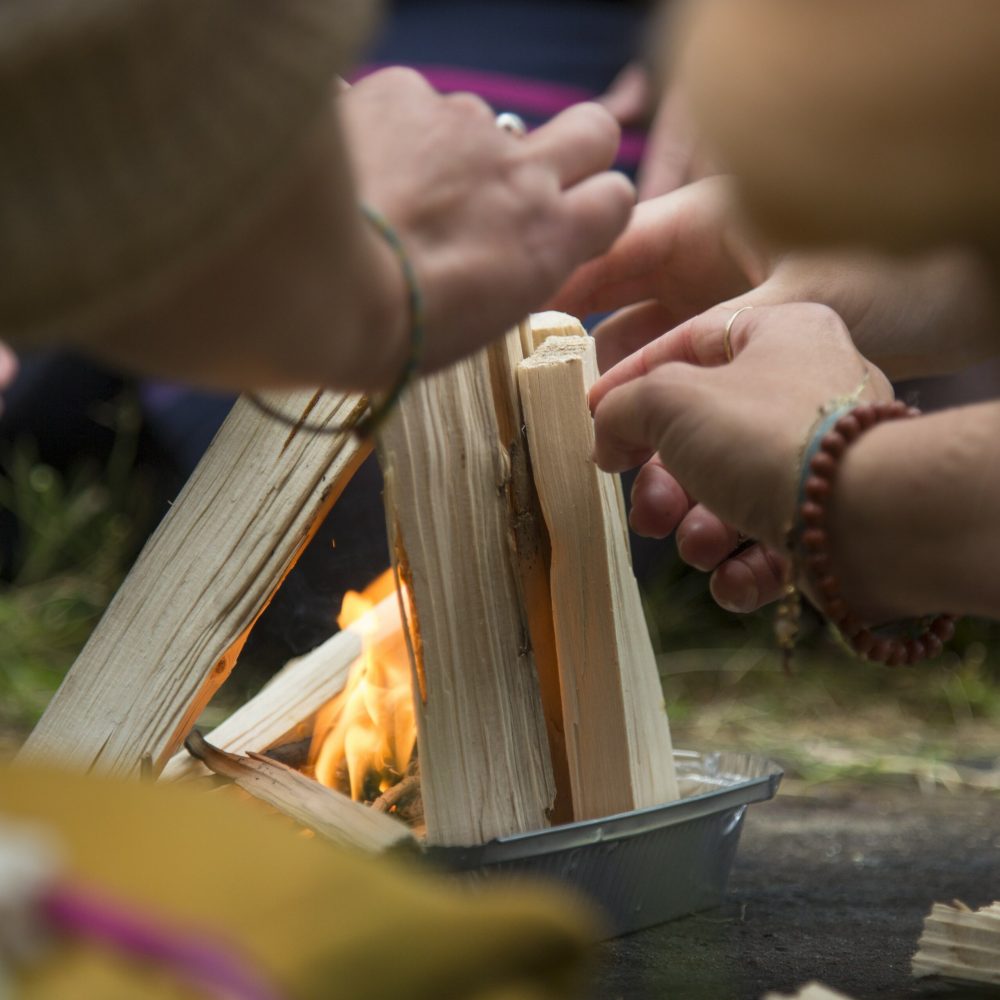 Man sieht im Rahmen des schamanischen Transformations-Retreats in Nahaufnahme, wie mehrere Hände kleine Holzstücke in einer Schale zu einem Feuer errichten, das Feuer brennt bereits in ersten zügen.