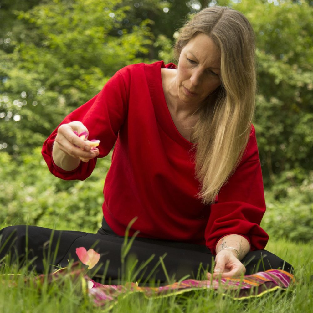 Man sieht Kerstin, eine Frau Mitte 40 mit blonden langen Haaren. Sie sitzt auf einer Wiese und lässt im Rahmen eines Stille-Retreats mit Sorgfalt Rosenblütenblätter auf ihr Medizinbündel fallen.