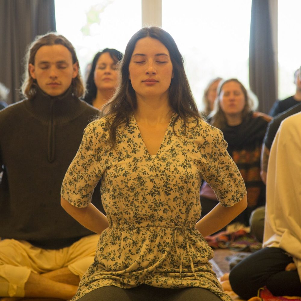 Man sieht eine Gruppe von Menschen verschiedenen Alters und Geschlechts, die mit geschlossenen Augen in die Meditation versunken sind. Auf dem Bild sind ca. 10 - 15 Personen zu sehen bzw. zu erahnen. Im Vordergrund des Bildes eine Frau in ihren 20ern, im Kleid mit langen dunklen Haaren, mit intensiver Ausstrahlung, tief eingetaucht im Rahmen eines Stille-Retreats.