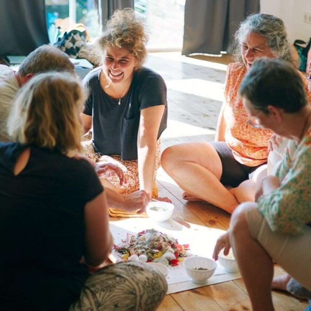 Man sieht im Rahmen des Transformations-Retreats sechs Menschen verschiedenen Alters in einem Kreis um eine Opfergabe herum sitzen, alles sind lachend im Gespräch.