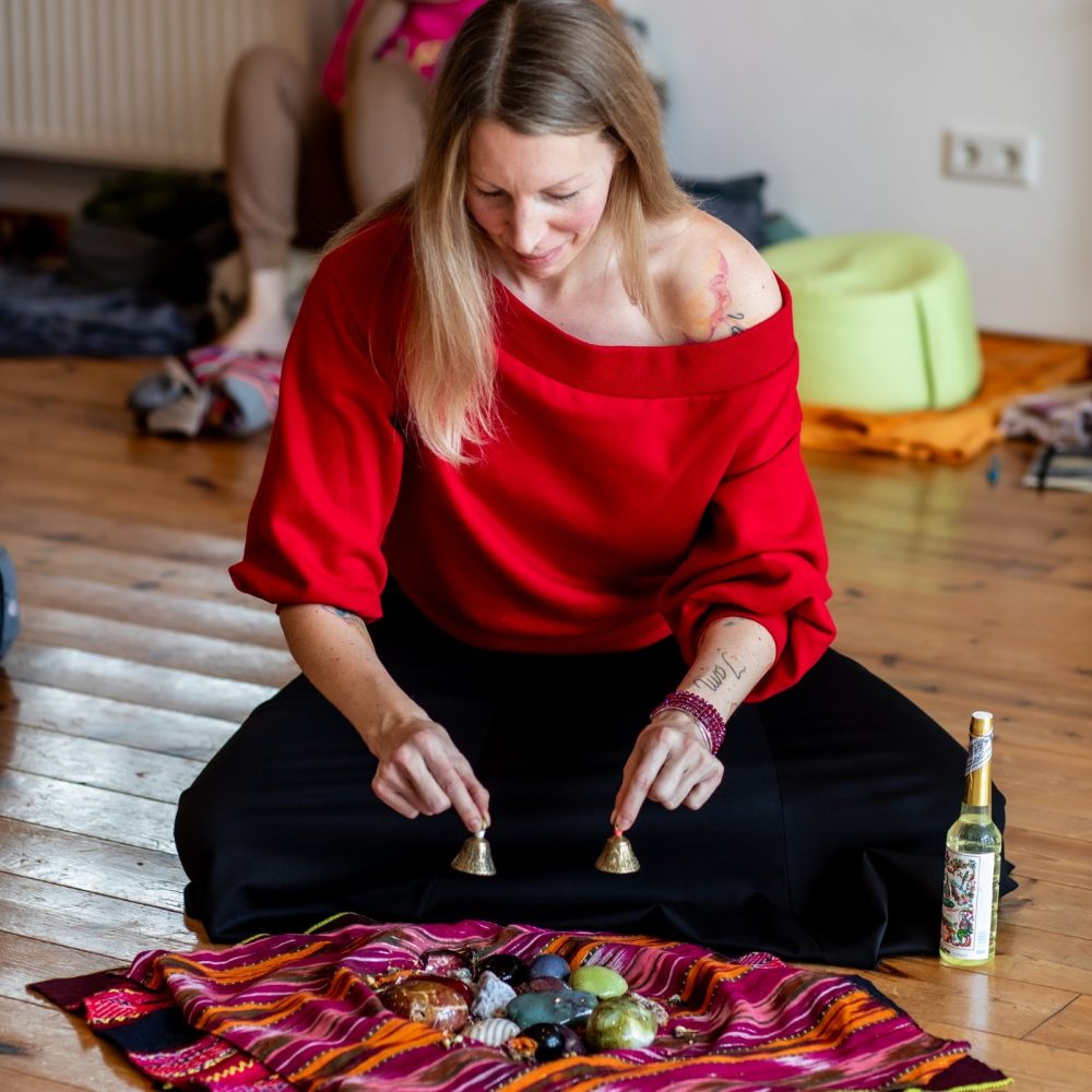 Man sieht Kerstin, eine Frau Mitte 40 mit blonden langen Haaren, mit langem Rock im Schneidersitz auf einem Holzboden sitzend mit Blick auf ihr geöffnetes Medizinbündel - auf dem Tuch ca. 13 verschiedenfarbige Steine - und mit goldenen Glöckchen in den Händen, die sie über die Steine schwenkt.